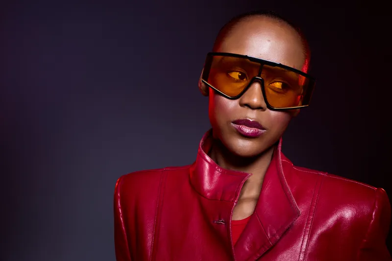 Stylish African woman with dark skin wearing oversized orange sunglasses and a red leather jacket looks away. Studio portrait with dramatic lighting and modern fashion concept.