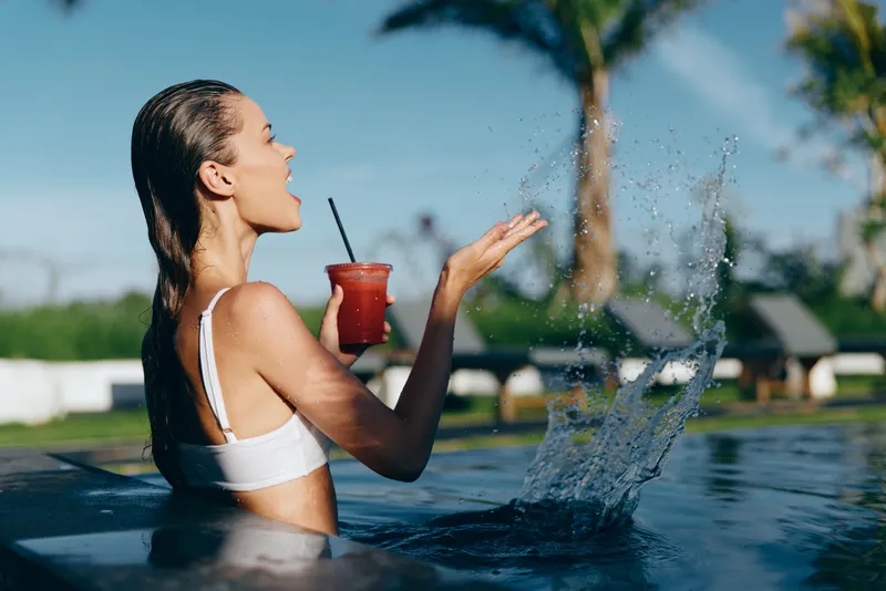 A woman in a white bikini enjoys a red beverage by a swimming pool, creating a playful splash as water droplets fly through the air, embodying carefree summer relaxation and resort style.
