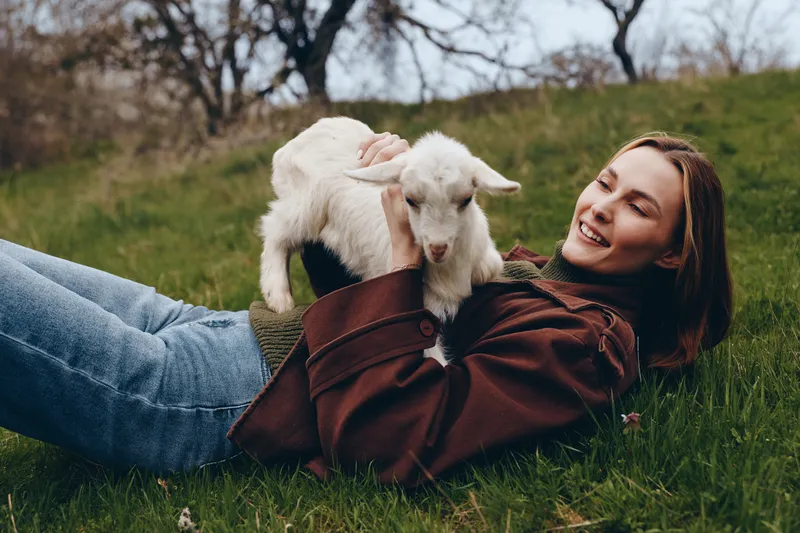 A woman lies on the grass, smiling as a goat kid sits on her chest, sharing a playful outdoor moment. This lifestyle scene captures companionship, rural charm, natural light, and simple joy in