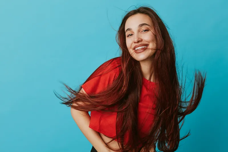 Young woman with long hair smiling and posing against blue background. Casual style, red top, happy expression, and dynamic hair movement in studio portrait.