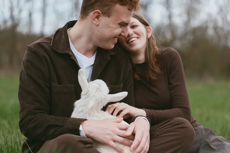 A joyful couple bonds with a playful goat kid in a sunlit field, sharing smiles and gentle touch. A candid lifestyle moment highlighting rural living, companionship, and animal affection.