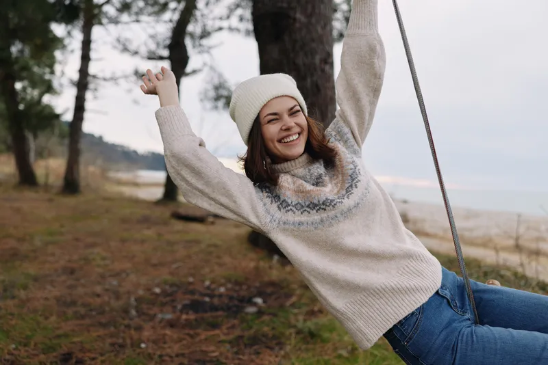 Woman on a swing outdoors in a cozy sweater and beanie, smiling as she enjoys a breezy day among trees and open space.