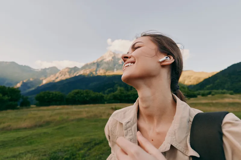 Smiling woman in a beige jacket outdoors, backpack on shoulder, enjoying nature amid green fields and towering mountains, soft sunlight highlights a relaxed lifestyle moment.