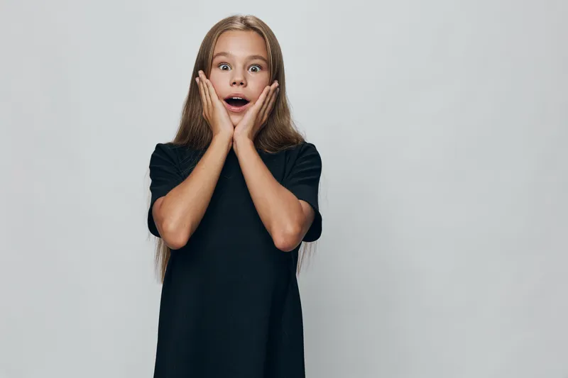 Girl with long hair wearing black dress showing surprise with open mouth and hands on cheeks standing in front of gray background. Child expression and emotion captured in studio.