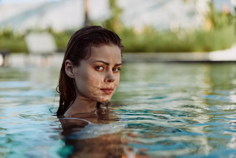 Young woman swimming in outdoor pool, looking over shoulder, wet hair, calm expression, natural light, lifestyle moment, water reflections, summer day