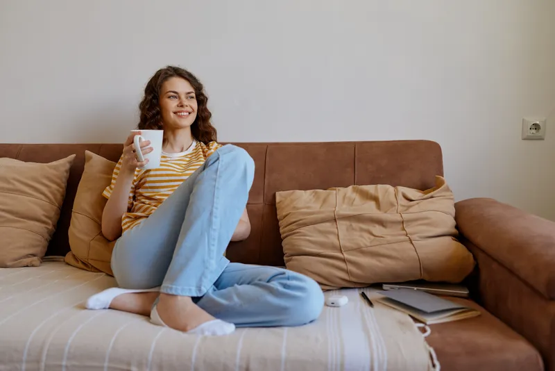 A relaxed woman lounges on a sofa at home, holding a mug and smiling in casual attire. A cozy living room scene portraying comfort, self care, and everyday home life.