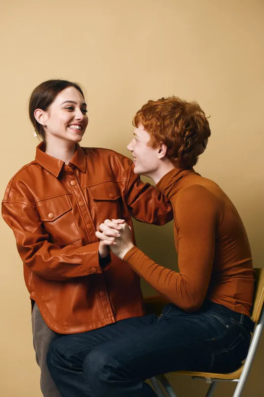 Two young adults sitting and holding hands smiling and looking at each other in warm clothing against plain background. Friendship, connection and happiness in casual indoor setting.