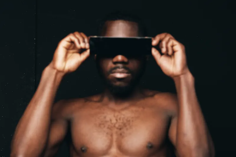 Creative conceptual portrait of African man with dark skin holding black object over eyes in visual style on black background. Artistic nude torso and focused expression in studio lighting.