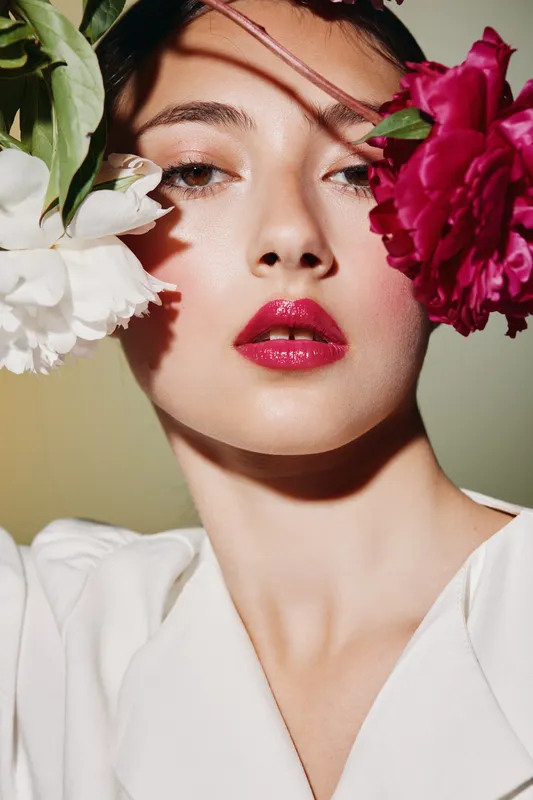 Portrait of a woman with glossy red lips and natural makeup framed by vibrant flowers. Soft light emphasizes freshness and fashion creativity. Beauty concept with floral details.
