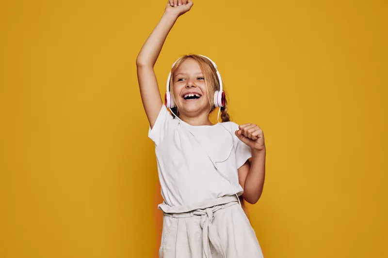 Happy girl with headphones dancing and enjoying music. She wears a white t-shirt and light shorts, smiling brightly against a vibrant yellow background. Concept for joy and music.