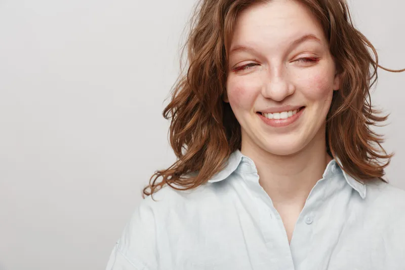Smiling young woman with wavy brown hair wearing a white shirt looks sideways with a playful expression on a plain light background. Concept of happiness, casual style, and natural beauty.