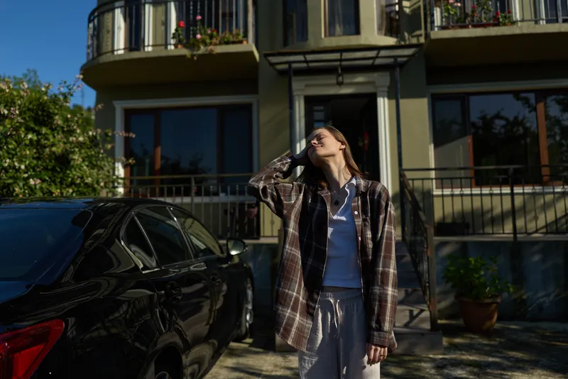 A woman in casual layers stands outside a modern building, shielding her eyes from bright sun. A parked car and greenery frame the scene, conveying everyday urban lifestyle and individual confidence