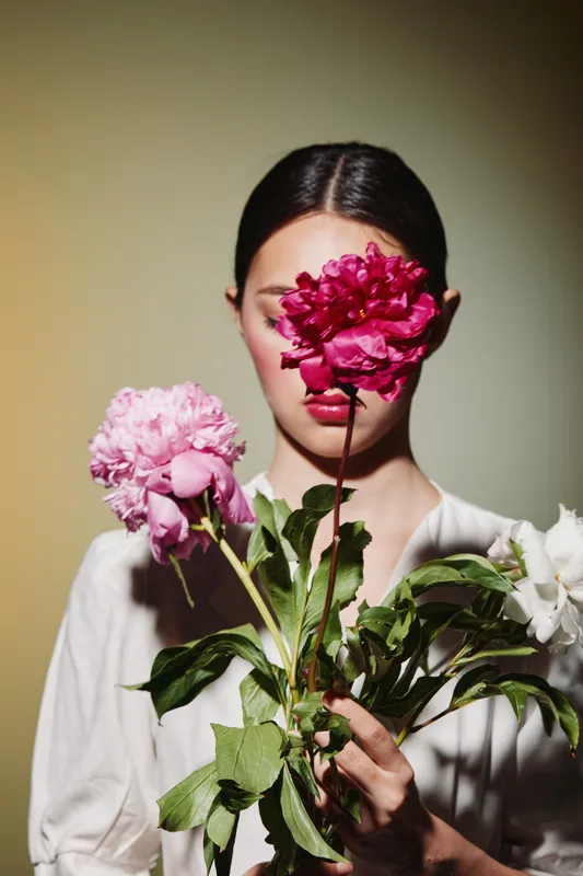 Young woman with clear skin holding vibrant pink peony flowers in front of her face. Minimal makeup, soft lighting, creative fashion editorial emphasizing natural beauty concept.