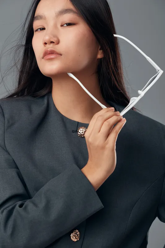 Young woman with straight black hair holding white glasses near face in studio portrait. Fashion model in dark outfit with confident expression and neutral gray background closeup.