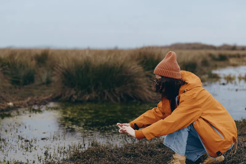 Woman in orange hat and yellow jacket walking outdoors by a river. Autumn nature lifestyle with wind, soft daylight, candid outdoor scene near water.