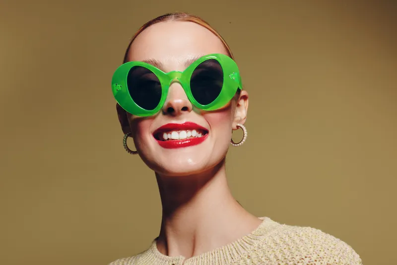 Smiling woman with bright red lipstick wears large green sunglasses and hoop earrings. She poses confidently against a plain beige background. Concept of fashion and style.