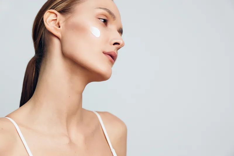 Young woman side portrait, beauty skincare cream on face cheek, bright indoor light, calm expression, minimal routine, clean skin texture, soft focus background, spa wellness mood