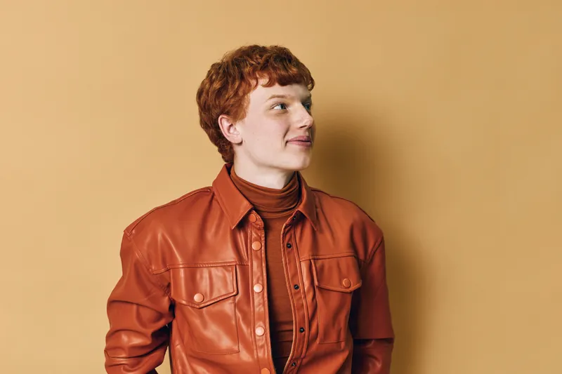 Young redhead man wearing leather jacket and turtleneck looking sideways with calm expression isolated on beige background. Portrait of confident casual male with curly hair in studio.