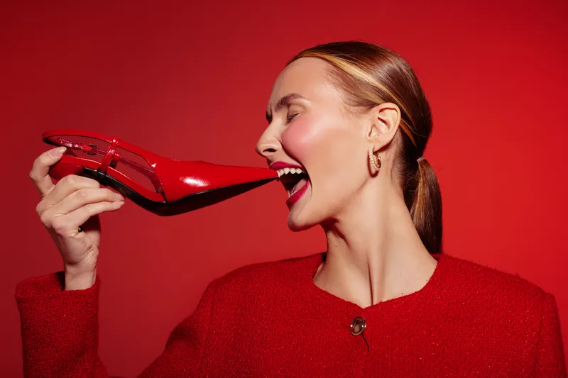 Woman in red jacket holds red high heel shoe near mouth on red background. Concept of fashion, style, and bold creative expression with vibrant color contrast.