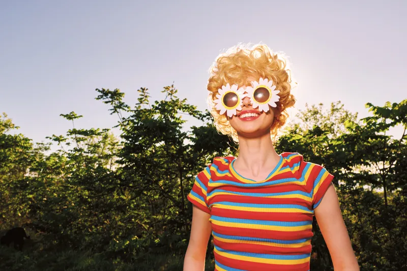 Woman outdoors, playful smile, striped colorful shirt, retro vibe, sunlit moment, light breeze, natural colors, greenery background, candid lifestyle shot, casual pose