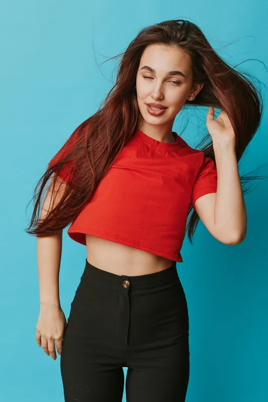 Young woman with long hair wearing red crop top and black pants posing with eyes closed against blue background. Casual fashion, portrait and beauty with natural look and movement.