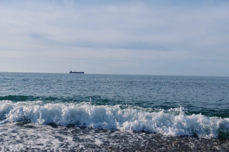 Sea, waves and distant ship on the horizon over calm waters, coastal shore with frothy surf, serene maritime scene suitable for travel, nature or oceanography themes in stock photography.