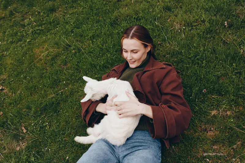 Cheerful woman outdoors, cradling a goat kid on a grassy field, wearing a warm jacket, sunlight glow, relaxed pose, candid moment, natural colors, pastoral lifestyle