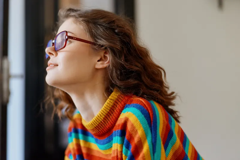 Young adult woman with curly brunette hair wears a colorful striped sweater and blue glasses as she gazes out a window, enjoying natural daylight in a bright indoor space, conveying calm and curiosity