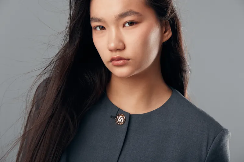 Young woman with long dark hair wearing a gray coat with a decorative button standing against a neutral background. Portrait, beauty, fashion and serious expression closeup.