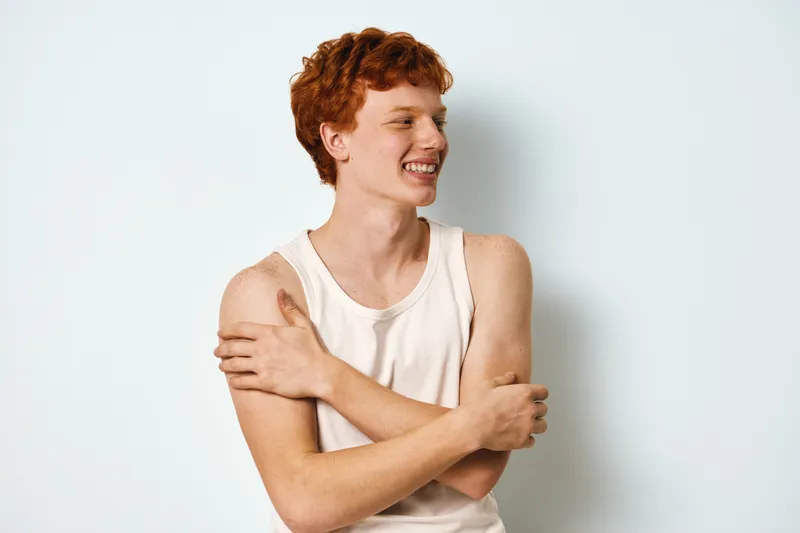 Young man with red hair in white tank top smiling and hugging himself on light background. Casual portrait of happy male expressing self care and confidence.