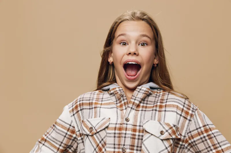 Girl with surprised expression and open mouth wearing plaid shirt posing on beige background. Child showing excitement and amazement in casual clothing in studio.