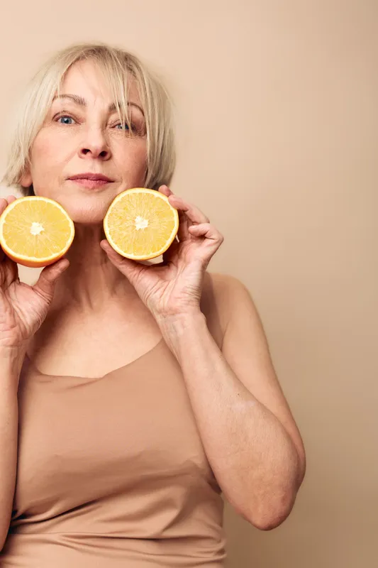 Mature woman with short blonde hair holds two orange halves near face on beige background. Concept for healthy lifestyle, natural beauty, skincare, and vitamin c benefits.