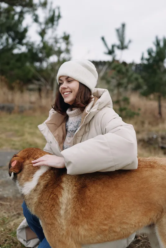 A woman in a warm beige coat and white beanie smiles while hugging a large brown dog in an open field during autumn.