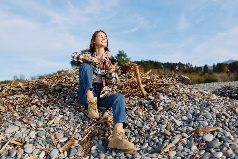 Woman enjoying outdoor casual moment on rocky pebbles, seated in denim and plaid shirt, boots on, relaxed in nature and landscape under clear sky, lifestyle scene of comfort