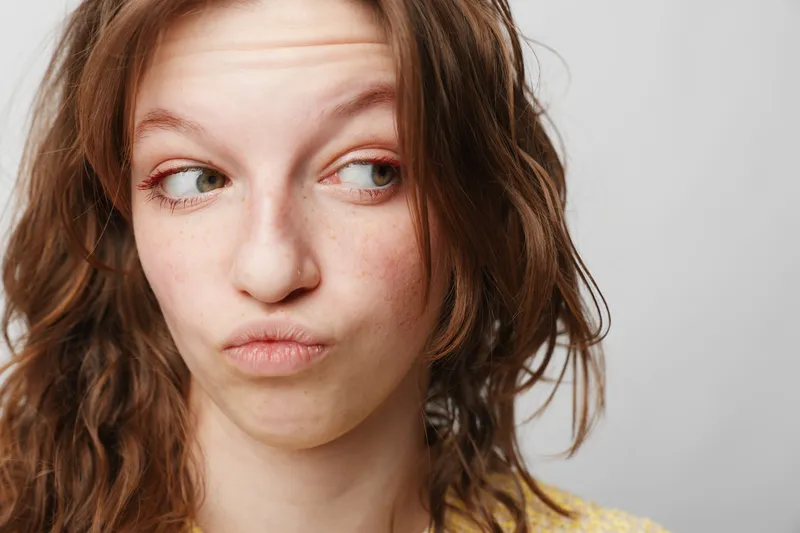 Close-up of a young woman with wavy hair making a skeptical facial expression looking sideways. Concept of doubt, curiosity, and human emotions in casual studio setting.