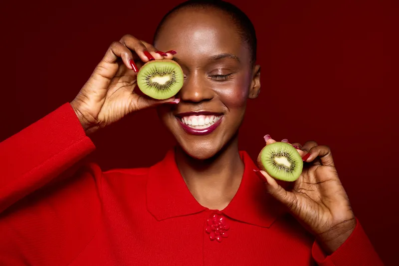 Smiling dark skinned African woman in red sweater holds two halves of kiwi fruit near her face against red background. Concept for healthy lifestyle, natural food, beauty and wellness.