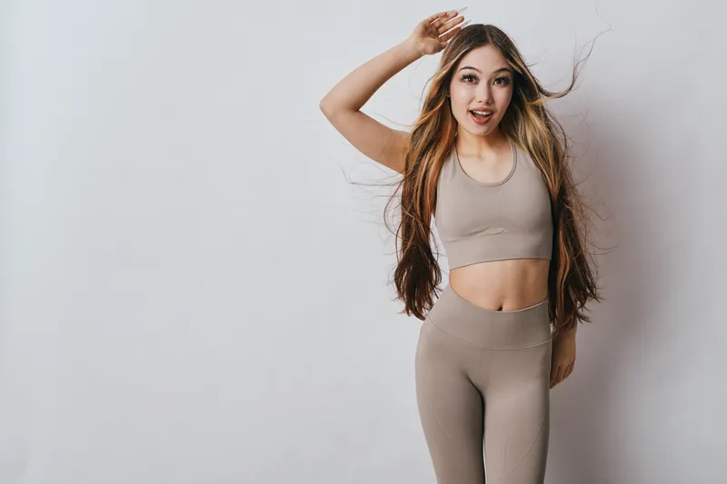 Young woman in beige activewear posing and smiling against white wall. Long hair flowing, fitness outfit with sleeveless top and matching leggings for workout or lifestyle.