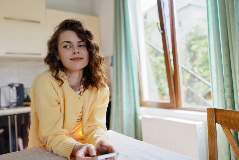 Casual woman indoors, seated at a table, wearing a yellow cardigan, looking to the side with a relaxed expression, sunlit home moment, soft natural light, warm colors Gentle breeze mood.