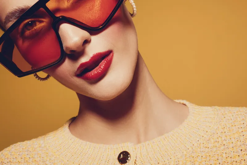 Close-up of stylish woman wearing large sunglasses and red lipstick against orange background. Fashion concept with trendy accessories and confident expression in studio lighting.