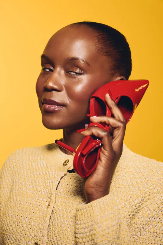Confident black woman with dark skin holds red high heel shoe near face. Yellow background and beige textured jacket create vibrant fashion concept with elegance and style focus.