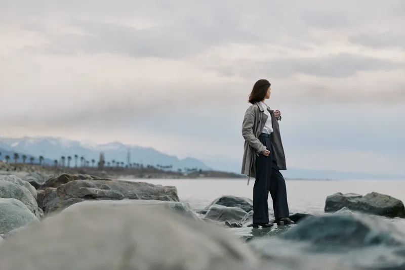 Young woman wearing coat stands on rocky shore looking at calm sea with mountains in background under cloudy sky. Concept for contemplation, nature, and outdoor lifestyle.