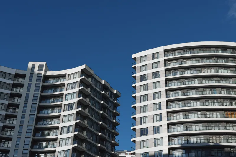Two modern residential buildings with curved balconies stand against a clear blue sky on a sunny day, showcasing contemporary architecture and urban living.