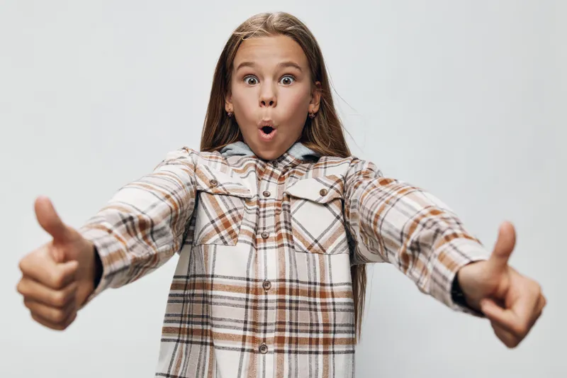 Girl in plaid shirt showing thumbs up gesture with surprised facial expression on white background. Child with long hair wearing casual clothing and positive hand signs.