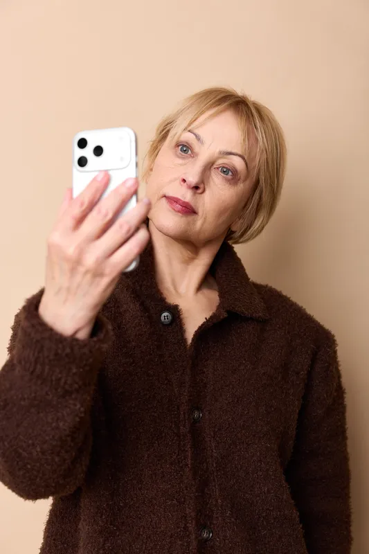 Older blonde woman in brown coat holds a smartphone for a selfie, portrait against beige wall, indoor communication for social media, thoughtful face and calm expression.