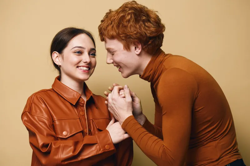 Young couple in casual clothing laughing and holding hands with happy expressions on neutral background for lifestyle and relationship concept.
