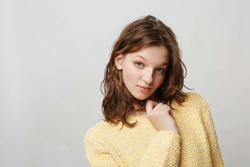 Young woman with wavy brown hair wearing a yellow knitted sweater poses against a plain light background. Natural beauty, casual style, and soft expression create a calm mood.