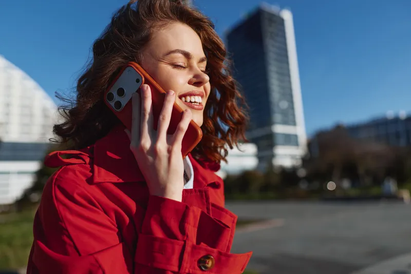 A woman in a red coat talks on a smartphone in a sunlit urban street, smiling with eyes closed, while tall buildings and clear blue sky create a vibrant city backdrop