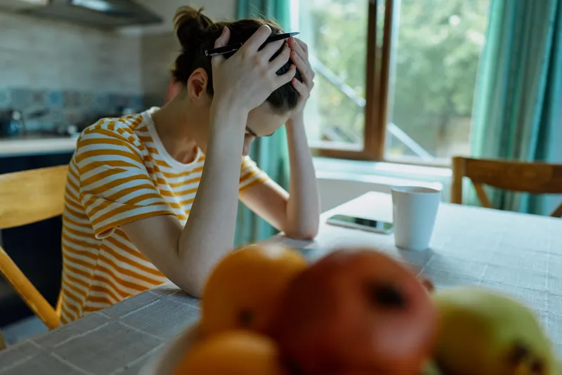 Casual woman at home, seated at a kitchen table, striped shirt, hands on head, worried expression, natural light, domestic setting, soft mood, everyday moment