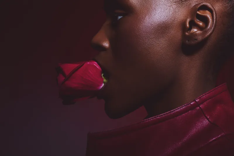 Profile of a dark skinned black woman holding a red rose in her mouth wearing a red leather jacket. Concept of beauty, sensuality, and fashion with vibrant colors in studio lighting.
