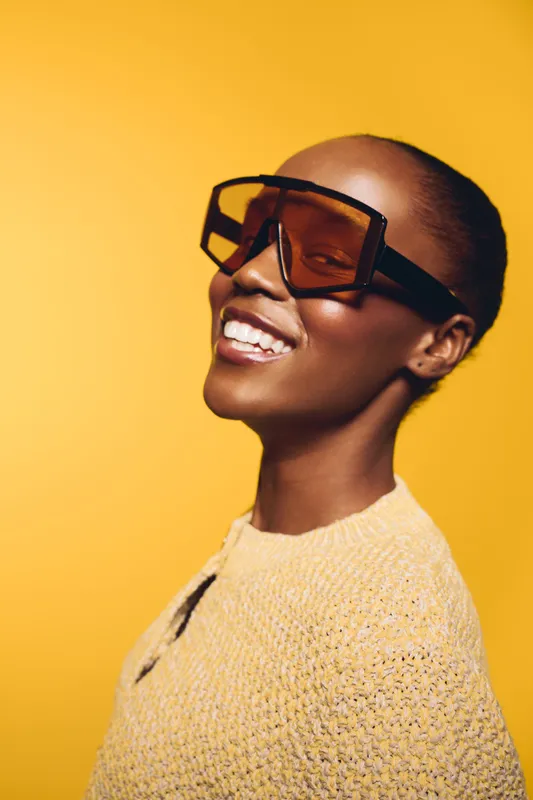 Smiling African American woman with dark skin wearing large sunglasses and a yellow sweater poses against a bright yellow background. Concept of fashion, confidence, and style.